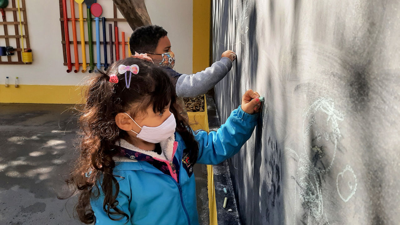 Imagem mostra menino e menina, usando uniforme azul, desenhando em parede pintada como lousa. 
Ao fundo, painel sonoro com canos coloridos.