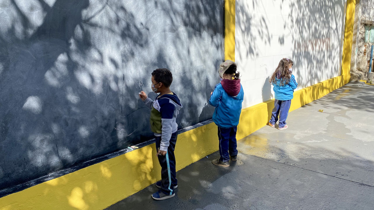 Imagem mostra painel de lousa e de azulejos brancos, três crianças desenham no espaço, uma sala de aula ao ar livre.
