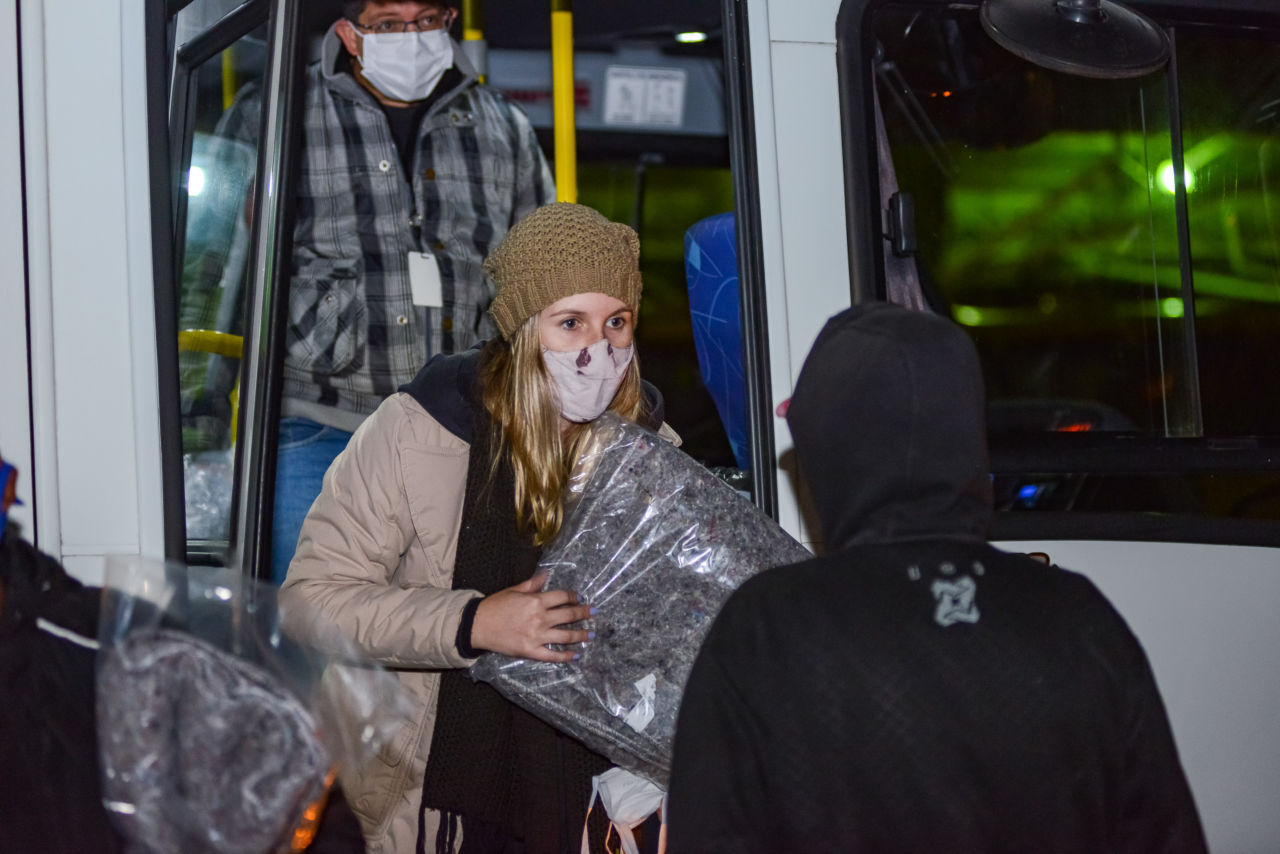 Mulher com gorro e máscara entrega pacote com cobertor a pessoa de costas com capuz, em frente a um veículo