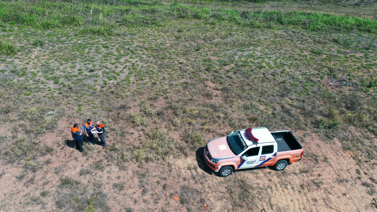 Foto aérea de três homens de jaleco verificando documento em terreno com vegetação rasteira e caminhonete adesivada