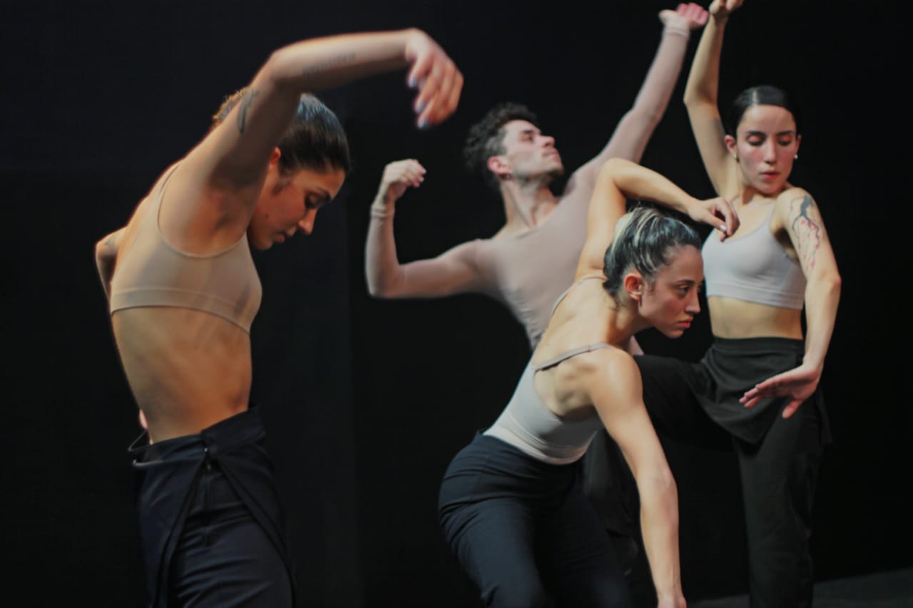 Bailarinos homens e mulheres, com calças pretas e parte superior da roupa com cor bege, fazendo movimentos de dança em frente a fundo preto