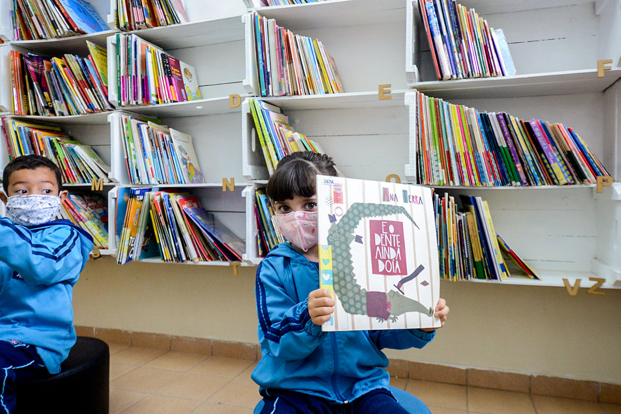 duas crianças, um menino e uma menina, com uniforme azul, estão sentados. A garota está com os cabelos presos e segura o livro "E o Dente ainda doia" nas mãos. 
