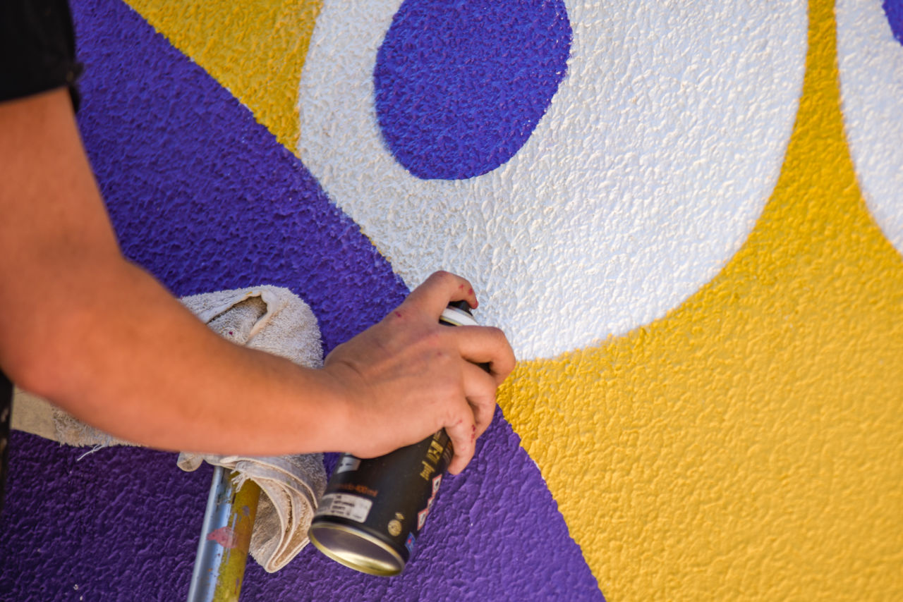 Detalhe de braço e mão segurando um spray de pintura, com um desenho coloridos de formar redondas na parede