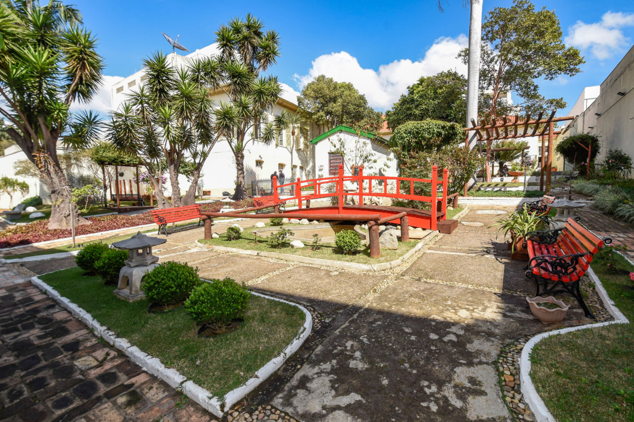 DESCRIÇÃO DA IMAGEM:
Jardim interno do Museu do Solar do Barão, pode-se ver uma construção de dois andares, com paredes brancas e janelas mais ao fundo. No centro da imagem uma ponte vermelha, com jardins e banco ao redor.