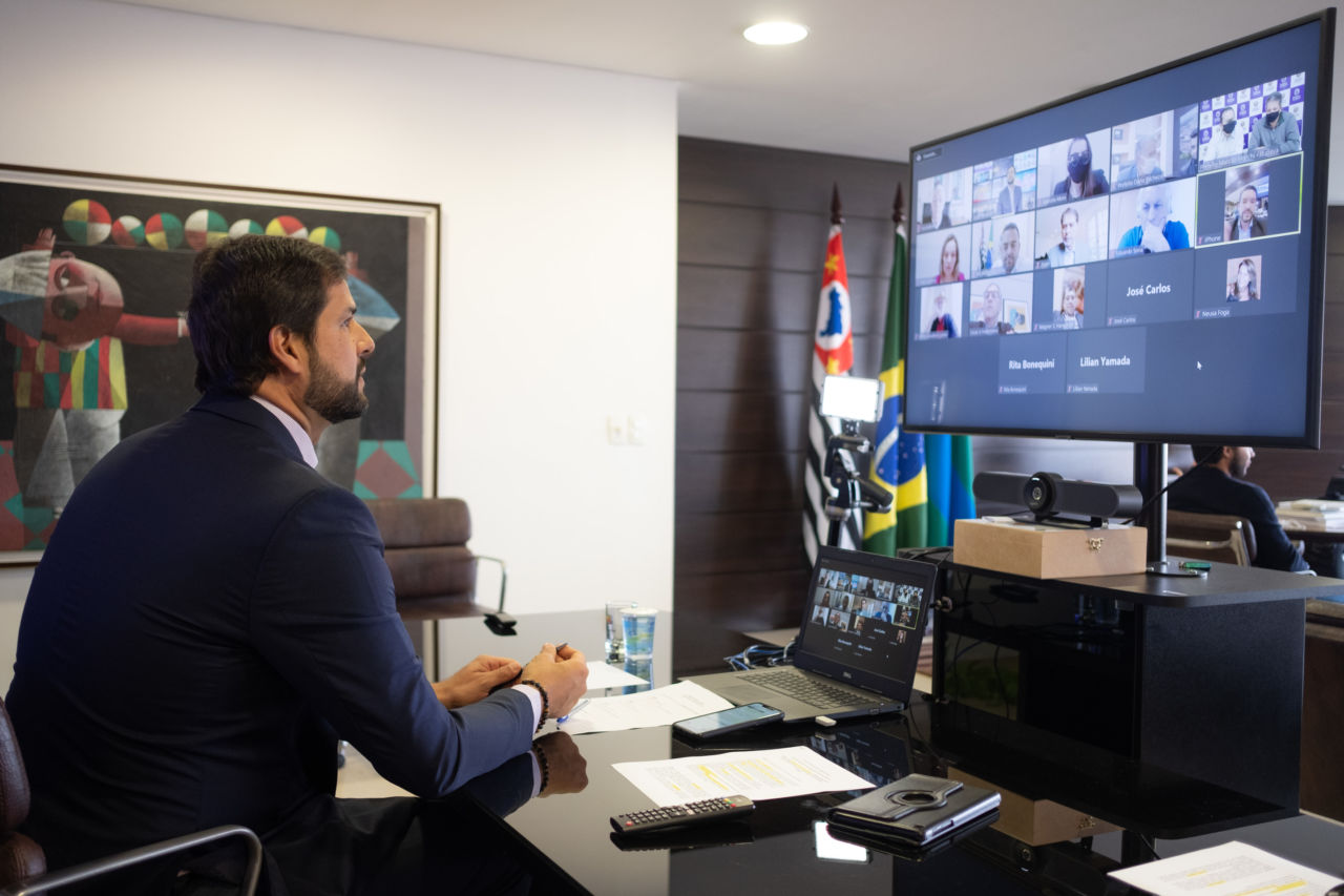 Prefeito Luiz Fernando (homem cabelos castanhos, barba, com terno azul) está em frente a um computador e uma tela de televisão, onde está uma reunião on-line, com a imagem dos diversos participantes. 