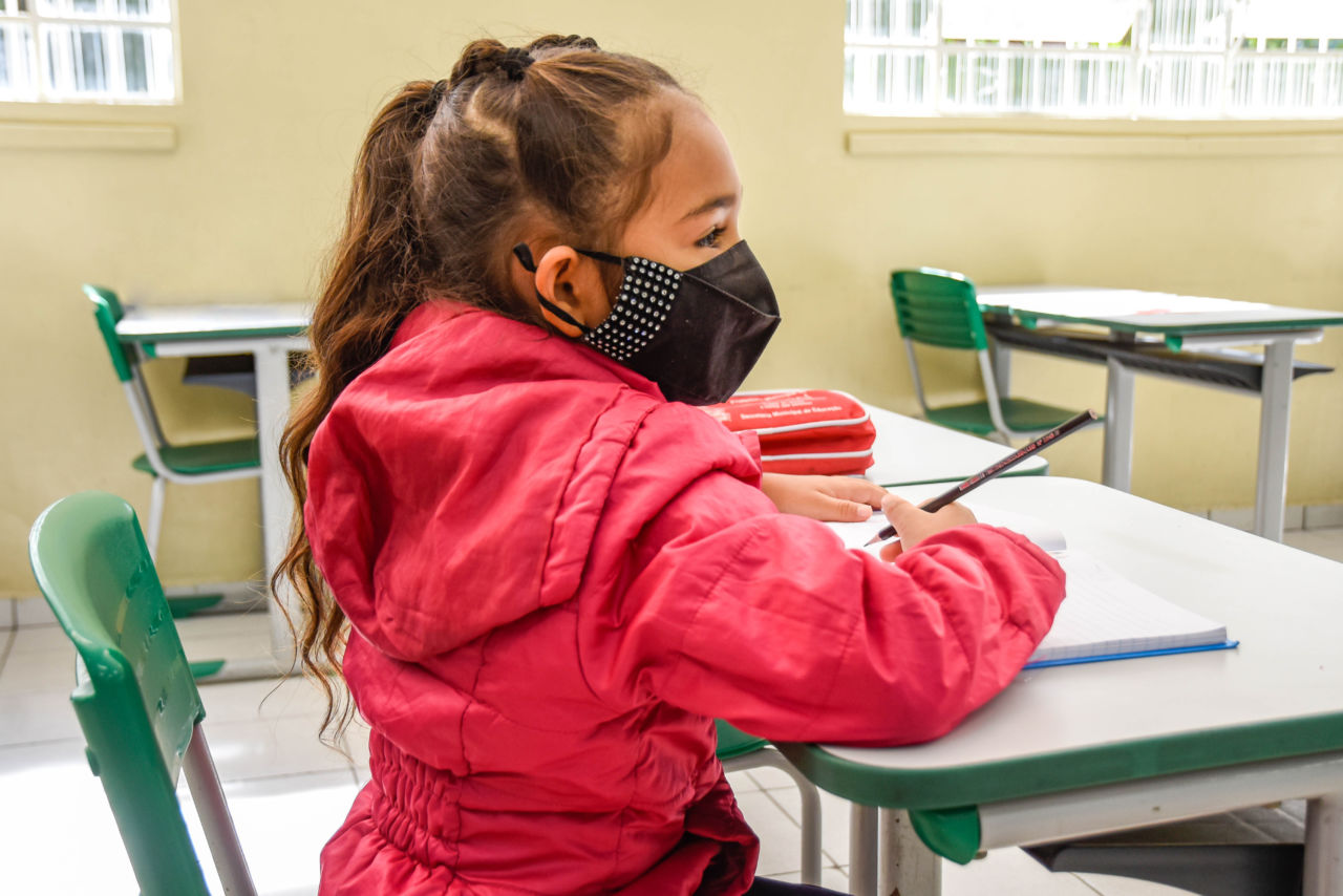 imagem mostra criança, com jaqueta rosa, sentada em carteira, de máscara preta. Ela olha para a frente e estão com caderno em cima da carteira e lápis em mãos