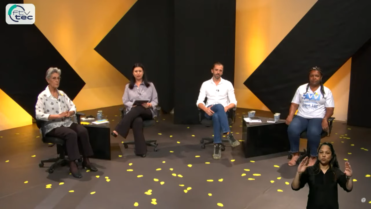 Três mulheres e um homem sentados em disposição semicircular, em estúdio de TV com faixas pretas e luzes amarelas em partes brancas da parede, com mulher fazendo sinais de libras no canto inferior direito da tela