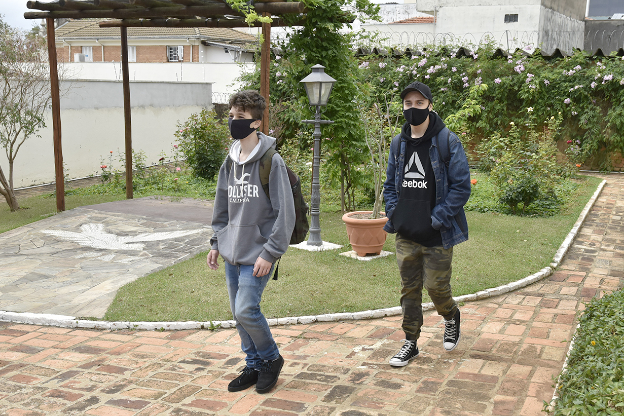 Dois moços, usando calças e roupas de frio e máscaras, caminhando em meio a jardim com flores e plantas