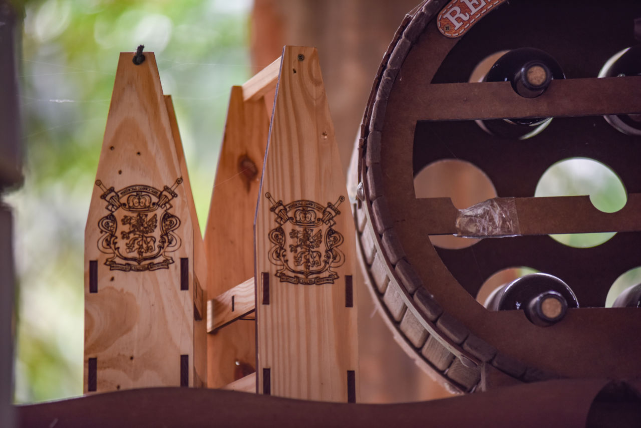 Imagem mostra adega de madeira, em formato de roda, com garrafas de vinhos deitadas, sendo possível ver a rolha de duas. Atrás há dois suportes de madeira para vinho, com brasão desenhado na lateral. 