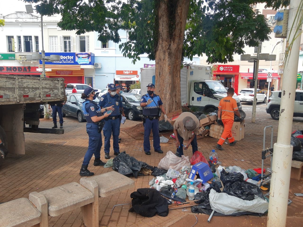 Praça da região central, com policiais e profissional da limpeza pública em torno de homem abaixado, usando boné, com lixo espalhado sobre o chão, com caçamba da caminhão ao lado.
