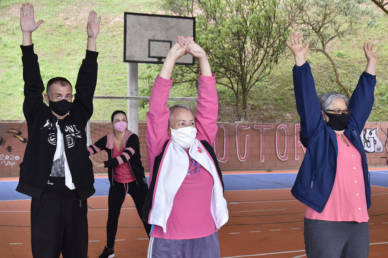 Três mulheres com blusas cor de rosa e um homem, todos usando máscaras, fazendo atividade física de alongamento dos braços, em quadra esportiva, com trave de futebol e aro de basquete ao fundo