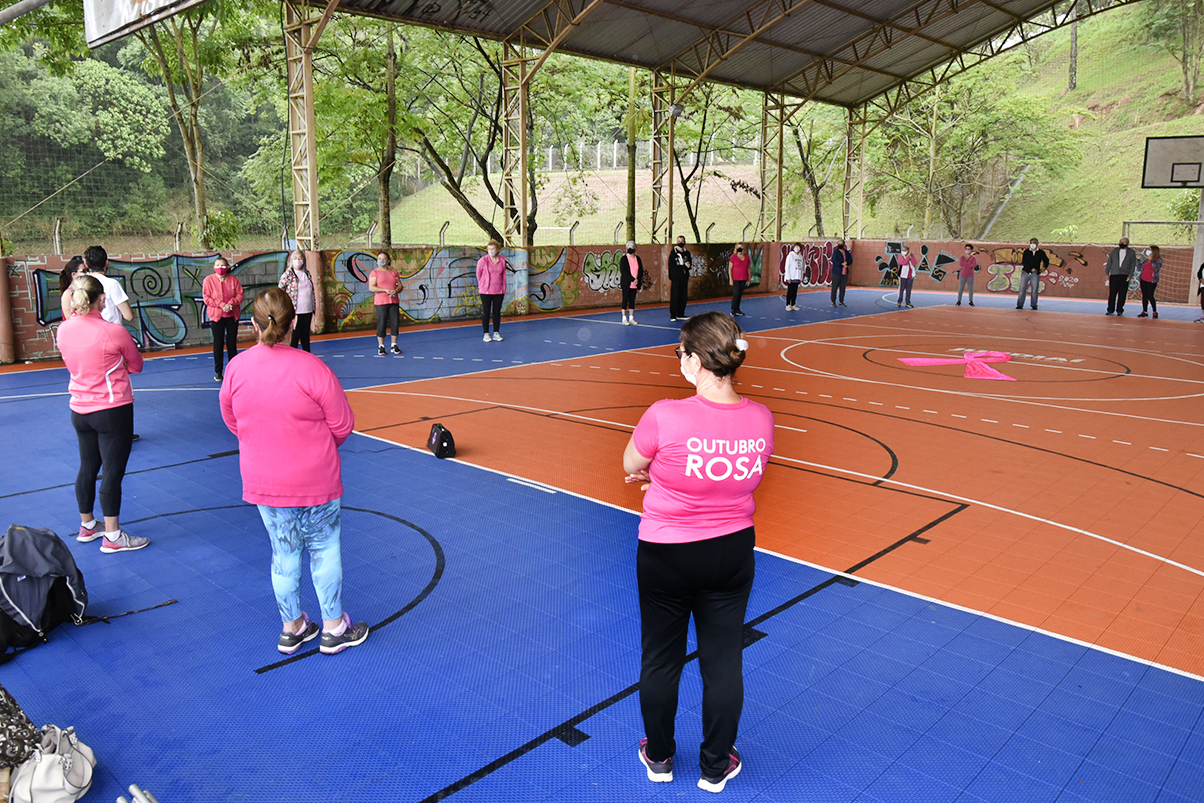 Homens e mulheres, todos usando máscaras e alguns deles com roupas cor de rosa, dispostos em formato circular, em ginásio esportivo coberto, com um grande laço de pano cor de rosa sobre o centro