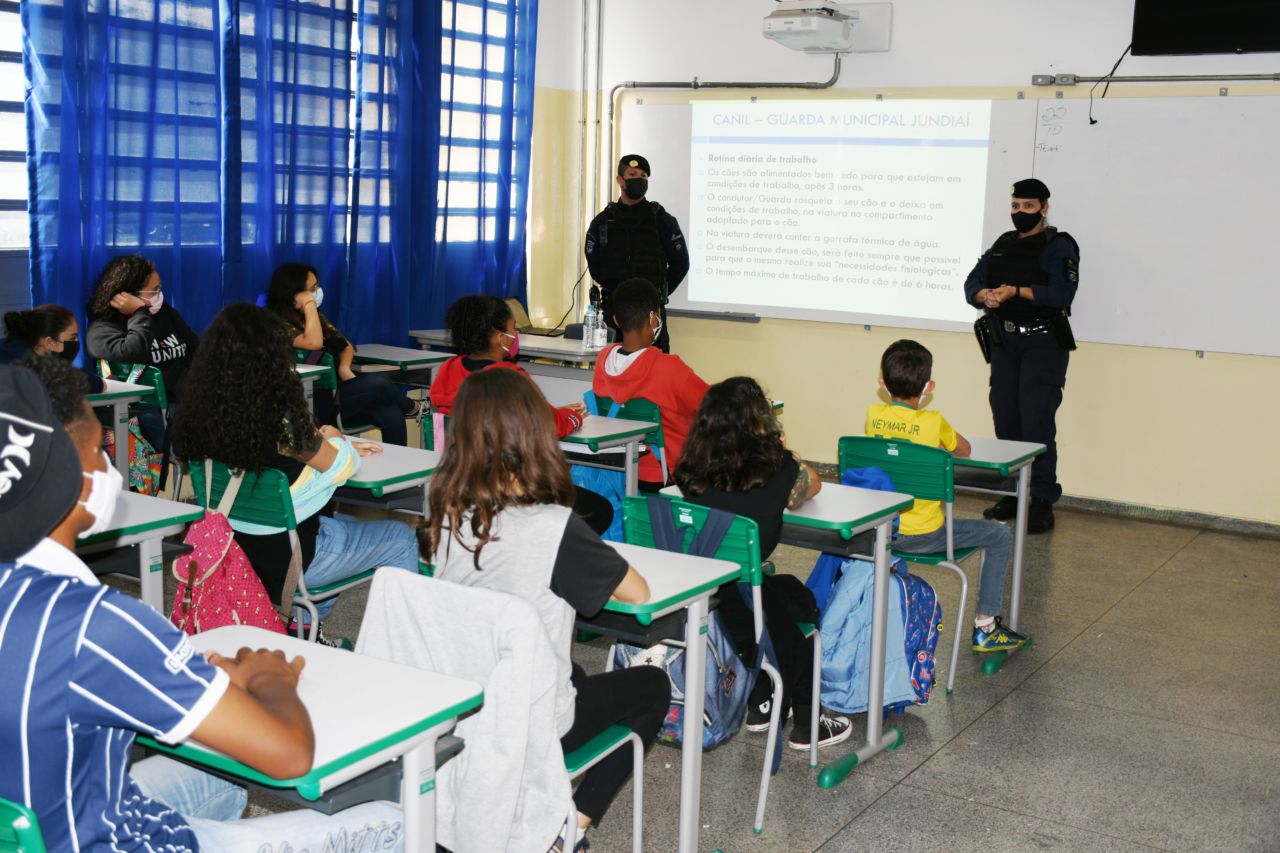 Um homem e uma mulher com uniforme policial, ambos usando máscara, fazem palestra para adolescentes sentados em cadeiras escolares dentro de sala de aula.