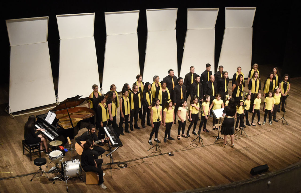 Homens, mulheres e crianças usando roupas pretas e amarelas, cantando, posicionados em fileiras sobre palco de teatro, onde também se encontra regente de costas e músicos tocando piano, violão e cajón