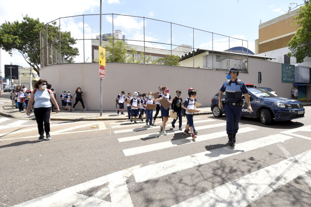 Crianças com uniformes escolares e máscaras, segurando pranchetas nas mãos, atravessam uma rua na faixa de pedestre, com homem com uniforme policial a pé e viatura policial na escolta