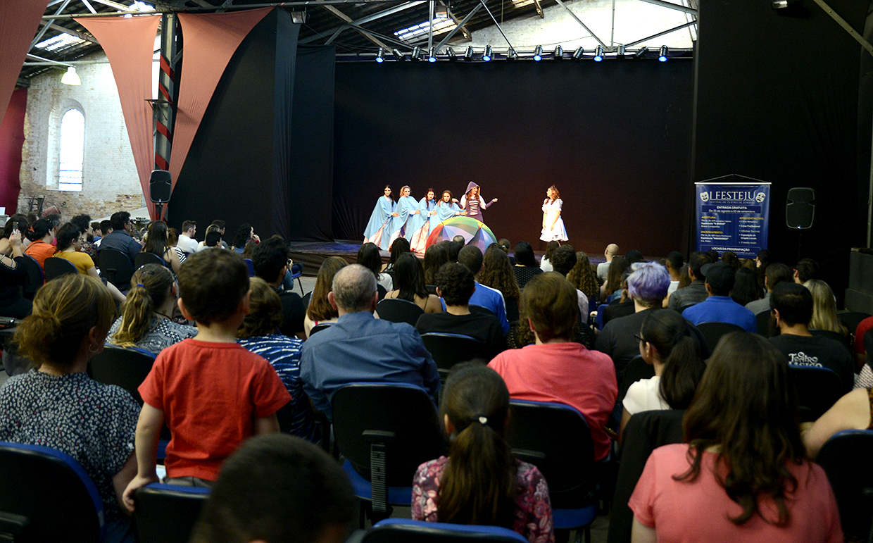 Sala de espetáculo, com pessoas sentadas em plateia, e grupo de teatro fazendo apresentação sobre palco usando capas de chuva e circunferência colorida