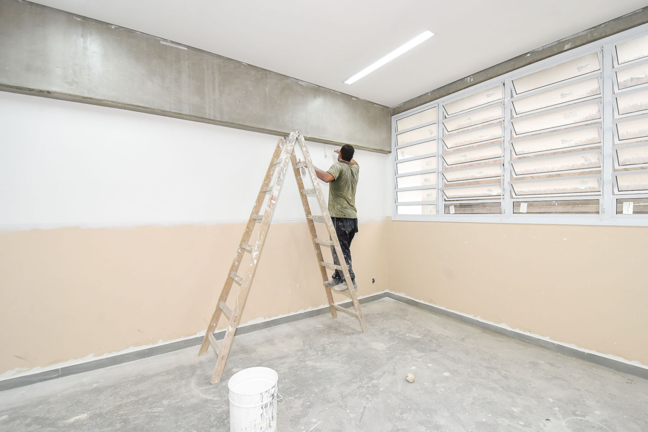 Homem está em uma escada de madeira, fazendo a pintura de uma parede. A parede está pintada metade branca e a outra bege.