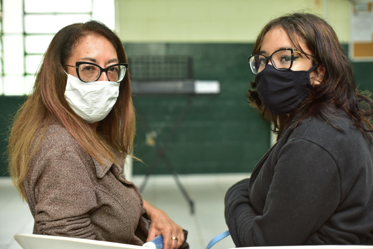 Duas mulheres de cabelos compridos, usando óculos, máscaras e roupas de frio, sentadas uma ao lado da outra, com o corpo retorcido para trás, olhando para a câmera.