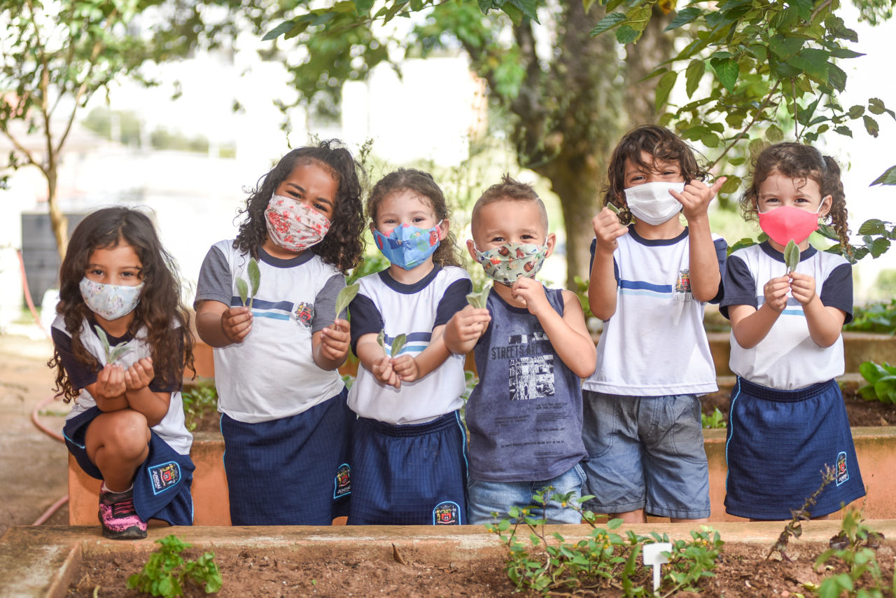 grupo de crianças está em uma horta com canteiros. Elas mostram folhinhas de plantas que seguram nas mãos. Todas usam máscara de proteção facial no rosto.