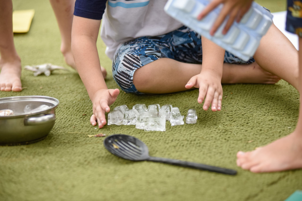 imagem mostra mãos e pernas de  criança brincando com gelos. Ela está sentada em cima de uma superfície que se assemelha a um tapete verde, que remete a uma grama.