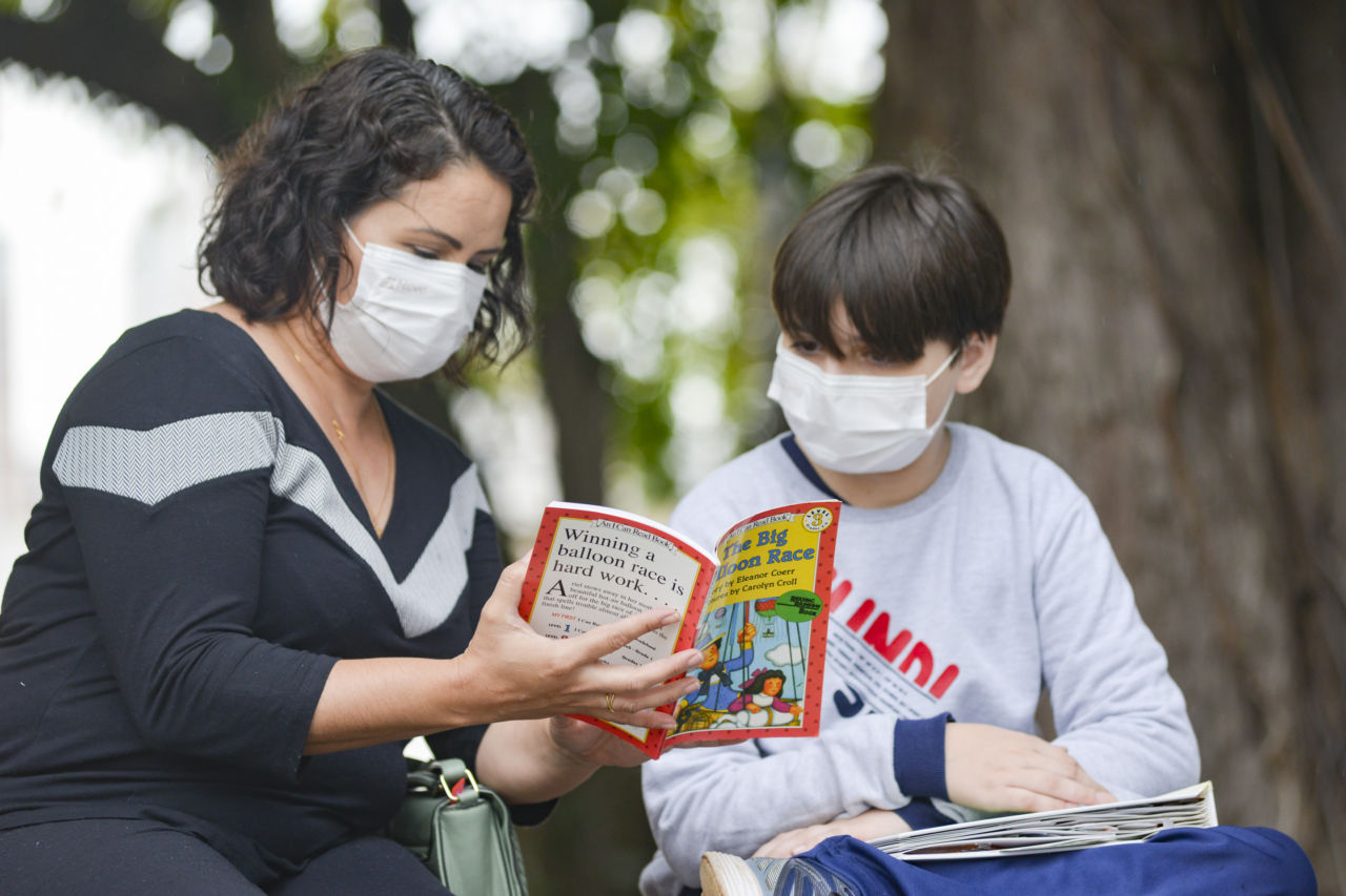 Mulher, com cabelos morenos, roupas pretas e máscara branca, está com livro com escritas em inglês nas mãos, ela mostra para um menino, com cabelos morenos, blusa cinza e máscara branca. Eles estão em um ambiente externo, com árvores ao fundo.