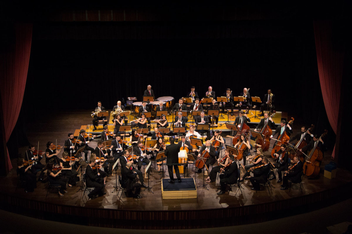 Imagem do alto de palco de teatro com orquestra se apresentando, com instrumentos de corda e de sopro