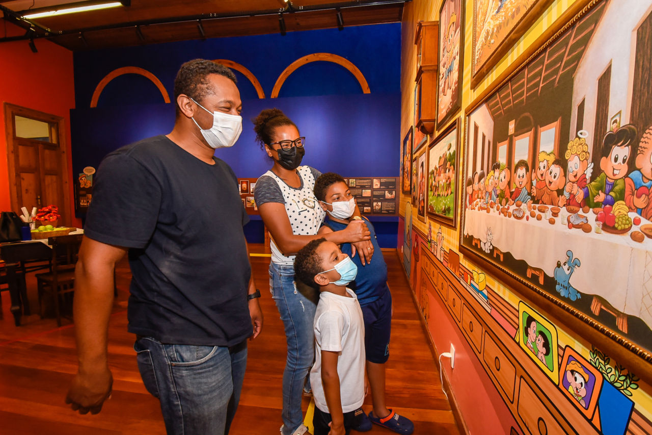 Casal e dois meninos usando máscaras conferindo as instalações de uma sala de exposições, com quadro de desenhos na parede