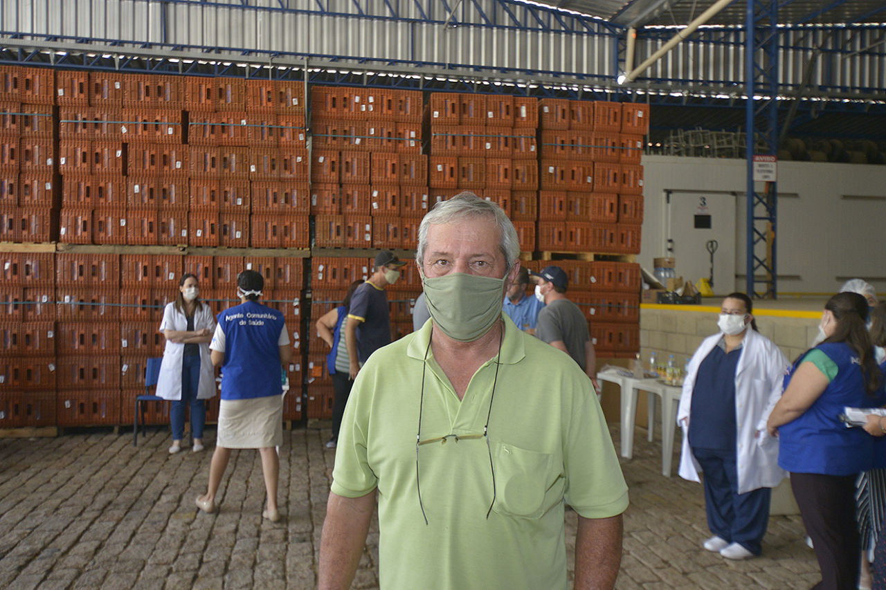 Homem de cabelos brancos, camiseta polo e máscara verdes, óculos com cordinha pendurada no pescoço. Atrás é possível ver outras pessoas e caixas de armazenamento de legumes/verduras na cor laranja. 