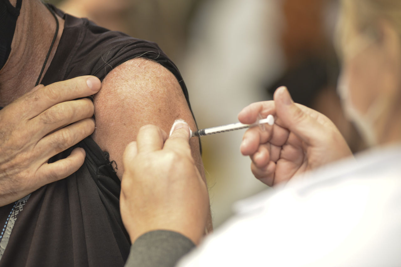 Aparece um braço de homem, que recebe aplicação de vacina por uma pessoa vestida de branco