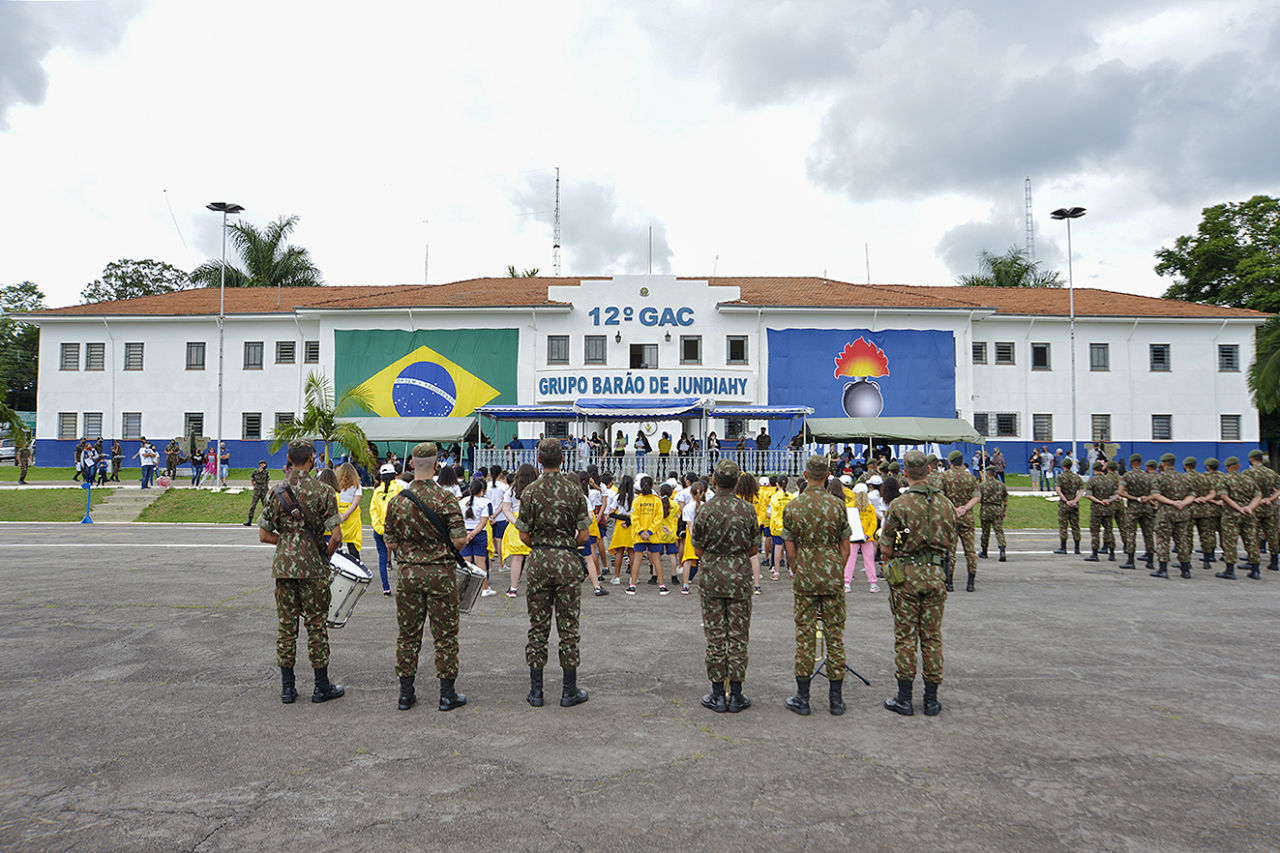 Imagem ampla com um prédio branco com janelas, o escrito "12° GAC" e "Grupo Barão de Jundiahy" com a bandeira do Brasil. De costas, estão os militares com uniformes camuflados, verde escuro. E na frente, as crianças com uniforme da ação amarelo e branco.