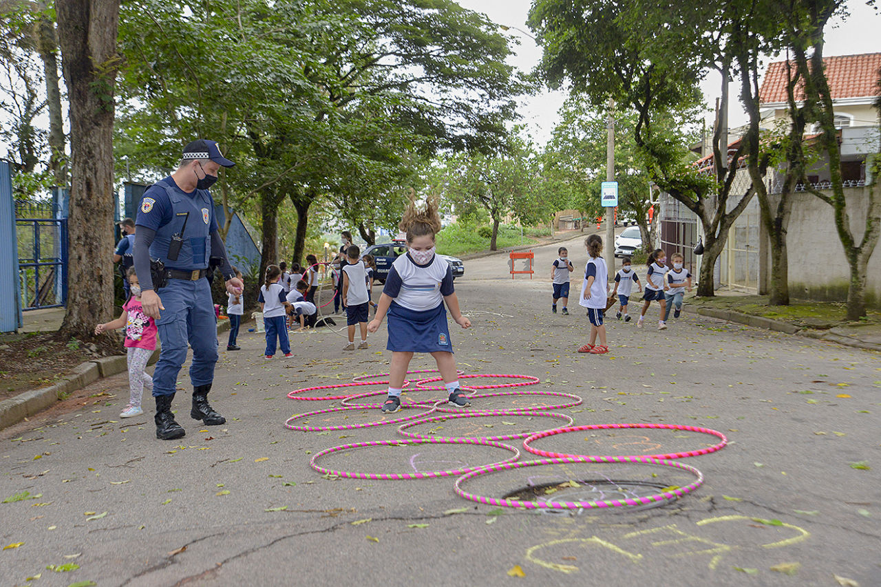Imagem ampla de uma rua com árvores com diversas crianças espalhadas de uniforme azul e branco. Encontra-se diversos bambolês rosas no chão com uma menina no ar, pulando dentro deles.
