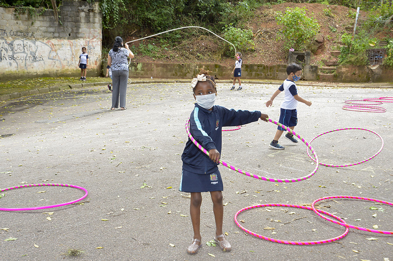 Uma aluna de uniforme azul e branco, e tiara com laço está com o bambolê rosa em volta do corpo, girando. Ao fundo há uma professora segurando a corda com uma aluna para uma criança pular e diversos bambolês espalhados pelo chão.