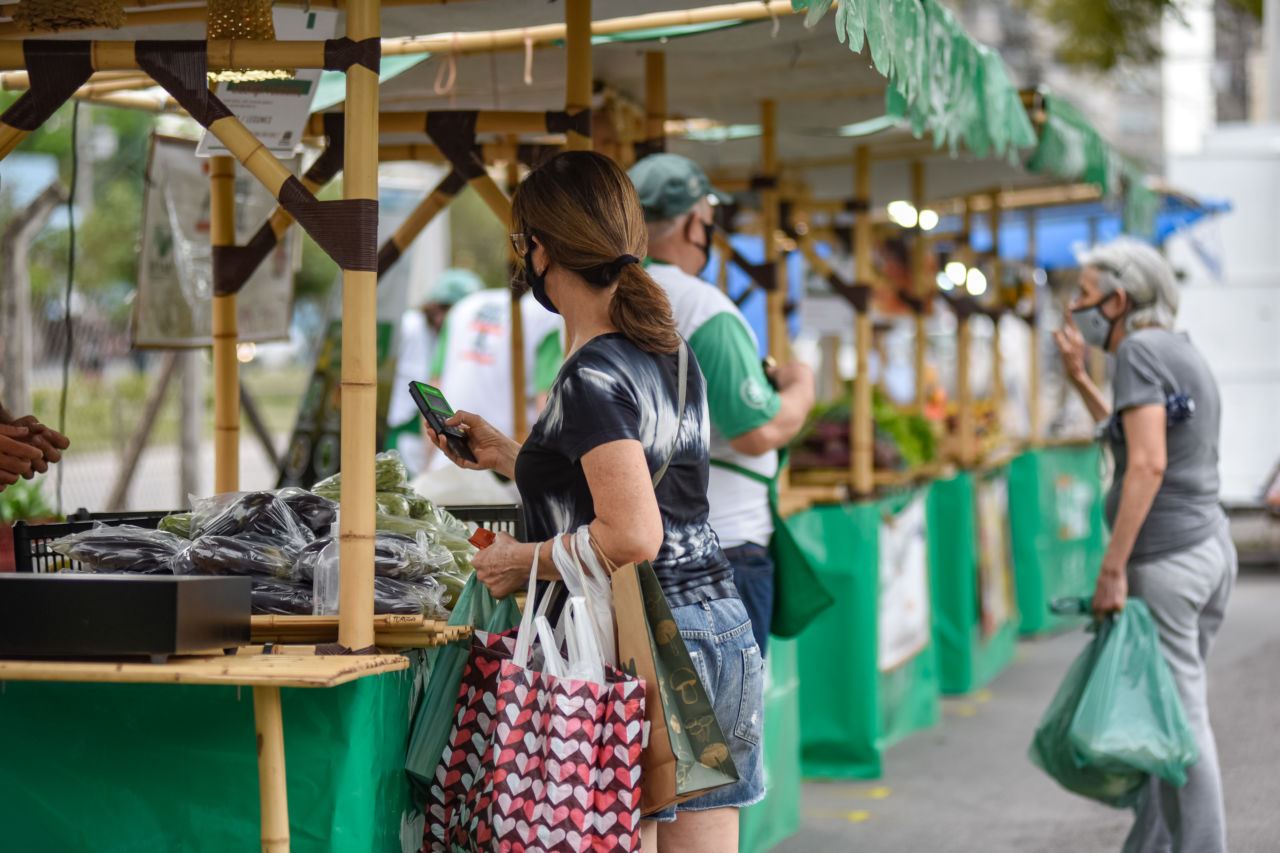 Imagem mostra feira com barracas com estrutura de bambu, tecido verde. Há pessoas fazendo compras de produtos. 