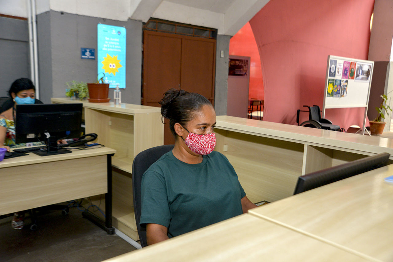 Mulher de máscara sentada atrás de balcão, com o olhar fico em tela de computador