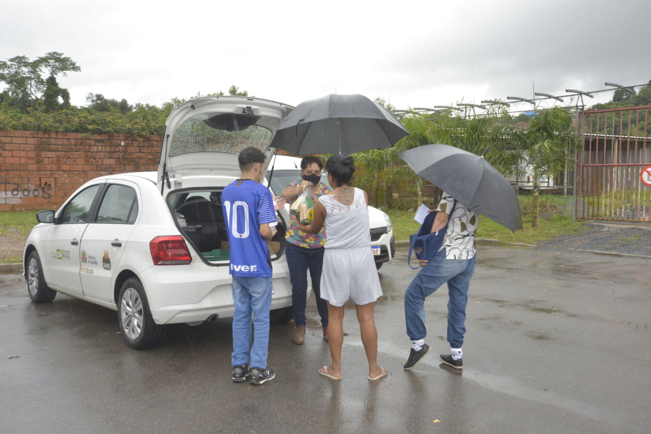 Ao lado de um carro branco da prefeitura, com o porta-malas aberto, um mulher faz a entrega de marmitex para três pessoas em área pública do Jardim das Tulipas