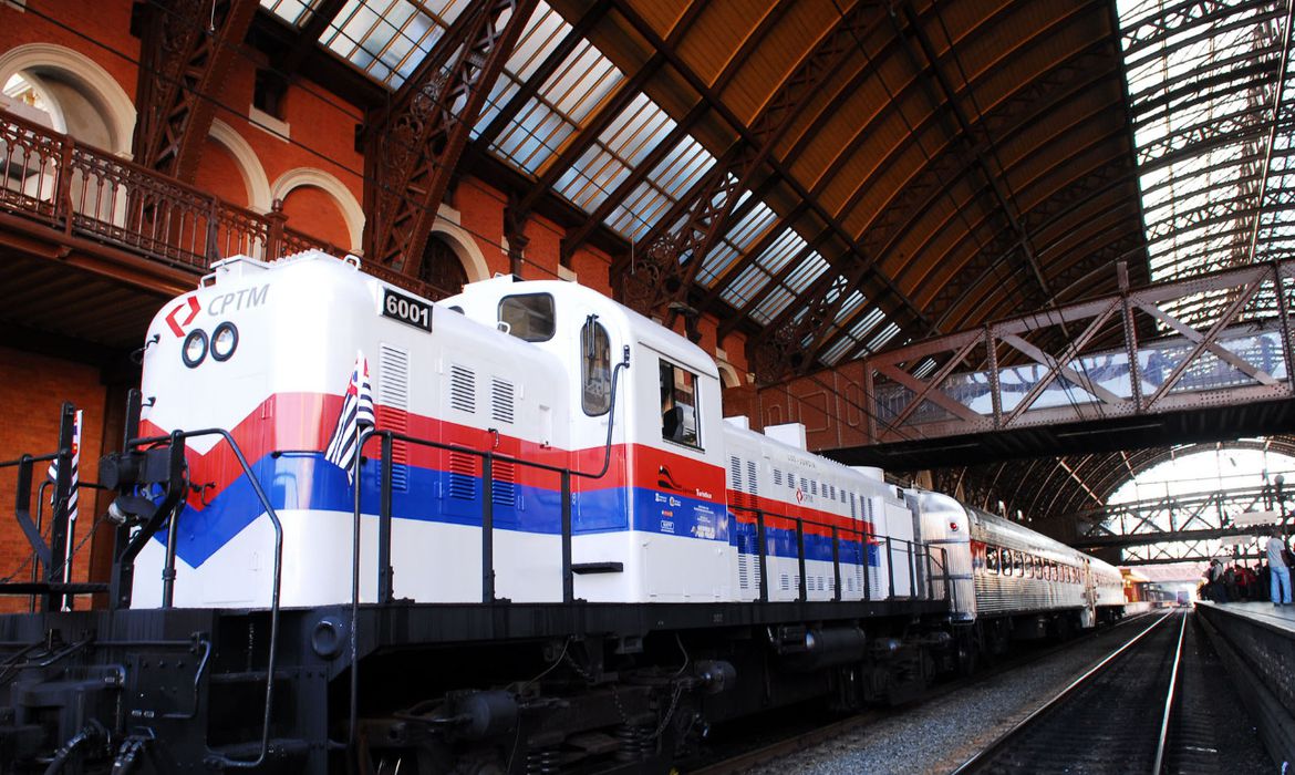 Em uma estação ferroviária, com detalhes em marrom, está estacionada uma locomotiva branca, com faixas azuis e vermelhas. Na frente lê-se CPTM e na lateral uma placa com o número 6001