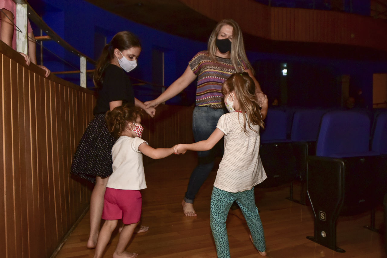 Uma mulher e três meninas dançam em roda, de mãos dadas, em plateia de teatro com poltronas ao lado