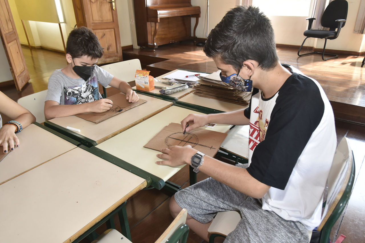 Dois meninos usando máscaras e sentados em lados opostos de cadeira escolar, fazendo desenhos com pedaços de carvão em pedaços de papelão