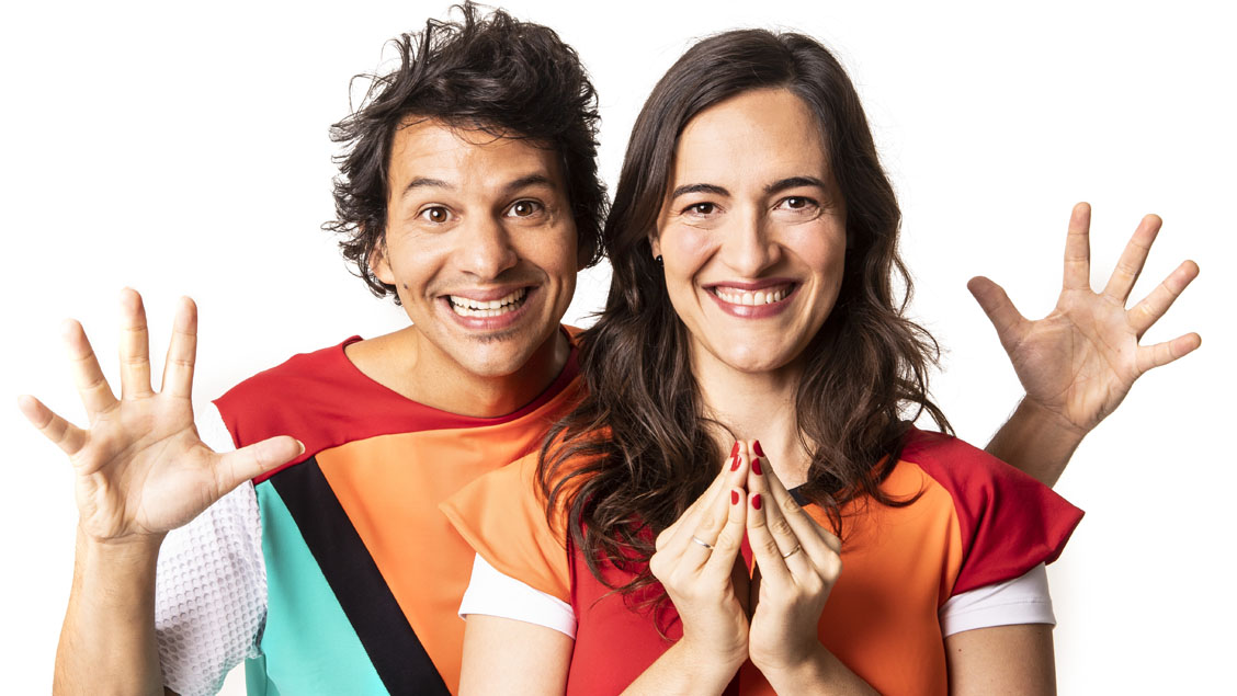 Homem de cabelo castanho curto e mulher de cabelo castanho comprido em foto posada com roupa colorida de formas geométricas, sorrindo e fazendo gestos com as mãos
