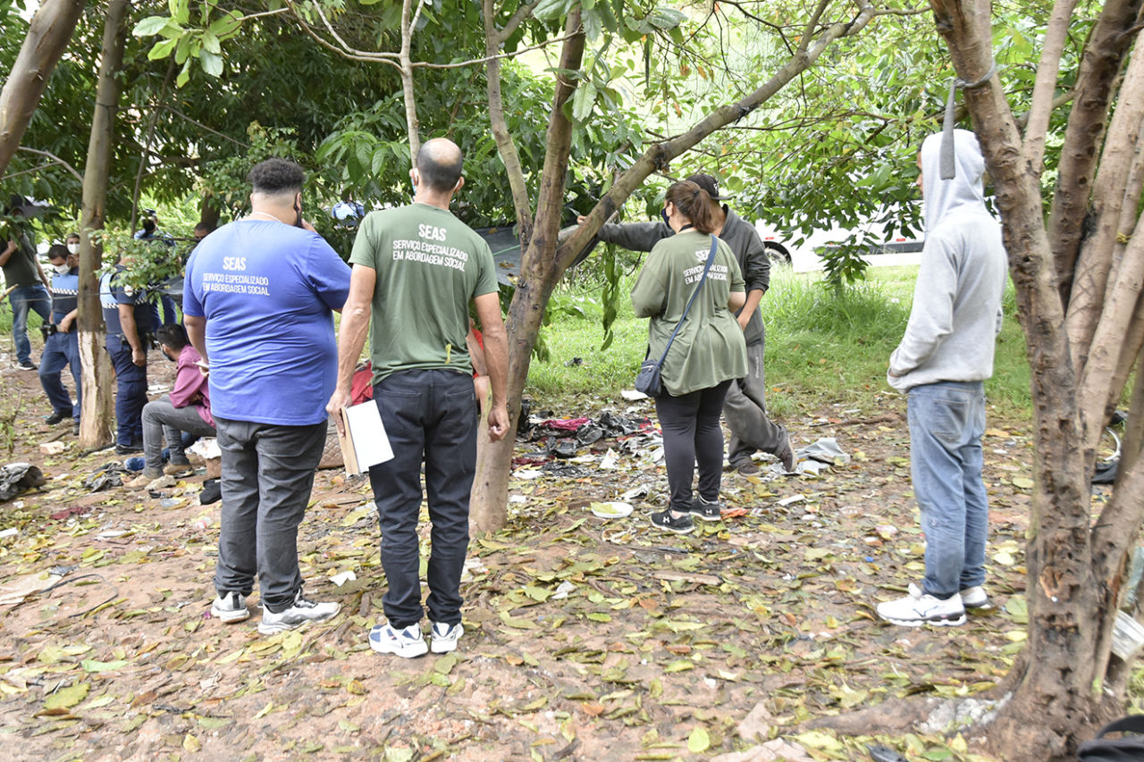 Pessoas com roupas uniformizadas de azul e verde, além de homens com uniformes de policiais., em jardim com árvores e lixo acumulado 