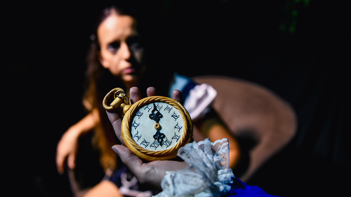Detalhe de mão com manga comprida azul e renda, segurando relógio de bolso com números romanos e marcação de horário às 07h08, com imagem embaçada de menina de vestido de babado ao fundo, sentada em cadeira, 