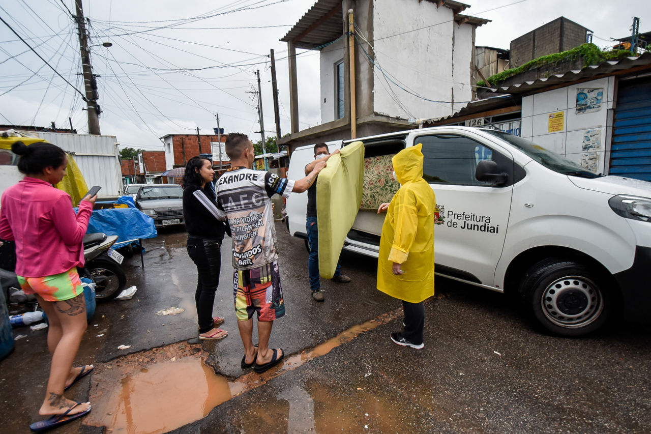 Duas pessoas com máscaras, uma delas com capa de chuva amarela, próximas a uma van, retiram colchões para entregar a moradores de área vulnerável