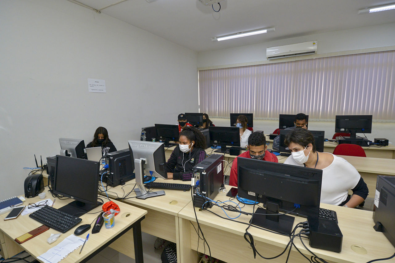 Sala de aula com diversos monitores de computadores em fileiras de mesas de madeira, com jovens usando máscaras