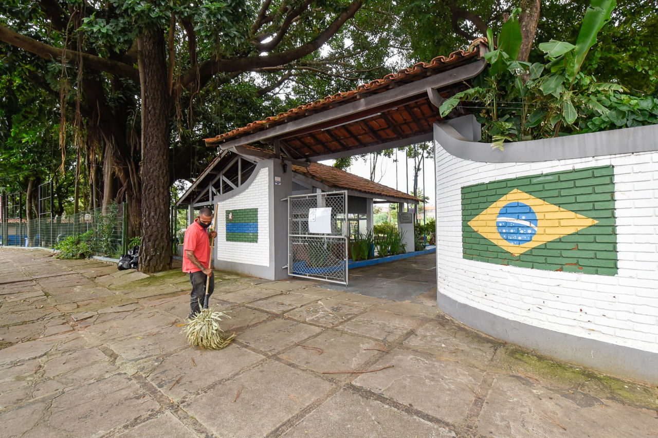 Homem de calça e máscara pretas e camisa de gola vermelha varre fachada de parque municipal com bandeiras de Jundiaí e do Brasil pintadas sobre a parede e árvores ao fundo