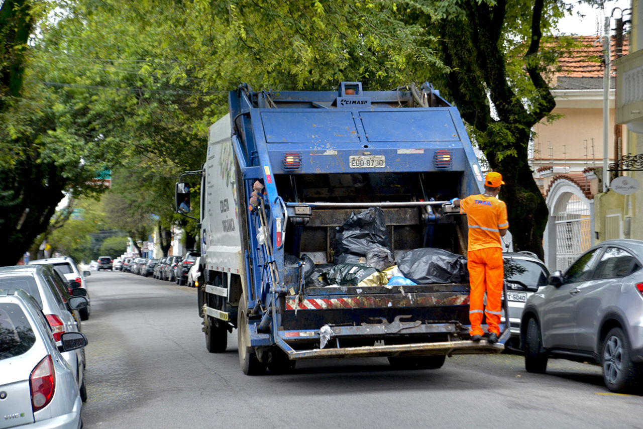 Descrição da imagem: caminhão de coleta de lixo em via, com coletor segurando nos apoios da caçamba, preparado para fazer a coleta dos sacos de lixo colocados pelos moradores
