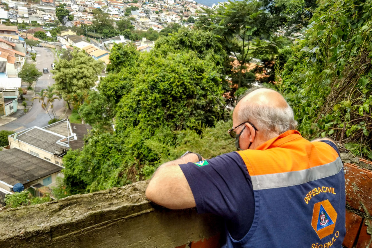 O assessor especial da Defesa Civil, João Osório Gimenez Germano, está encostado no muro, olhando para baixo, para o morro em que ocorreu deslizamento de terra; ao fundo é possível ver as árvores do local e o teto de casas