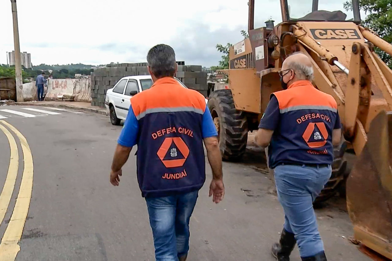 Com coletes laranjas, técnicos da Defesa Civil caminham por rua do Jardim São Camilo, ao lado de um trator
