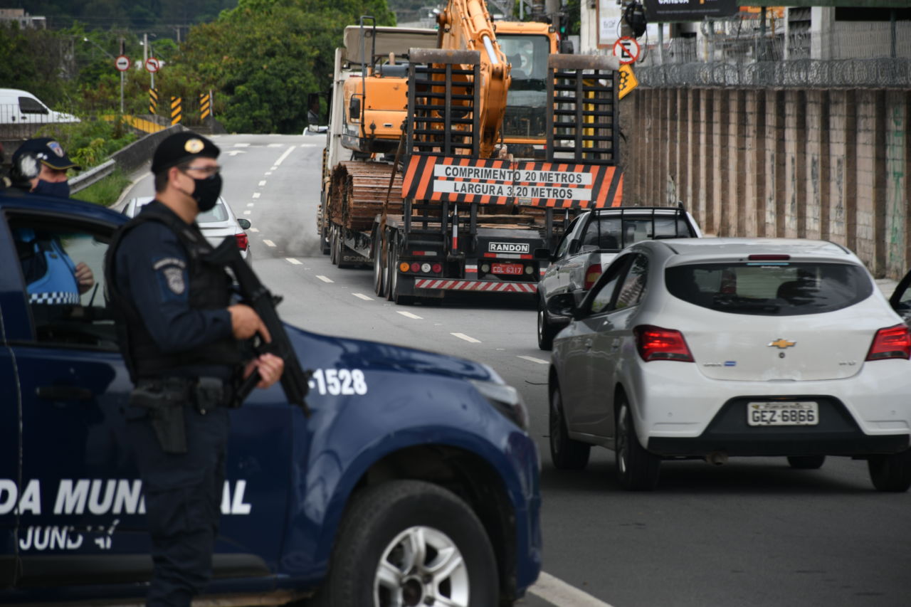 Ao lado do veículo, com uma metralhadora em mãos, guarda municipal observa o trânsito na avenida Frederico Ozanam; um carro branco e um caminhão passam pelo local