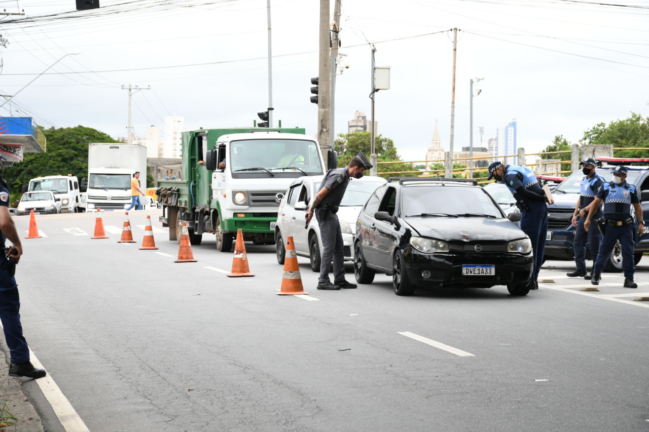 Policial militar observa interior de veículo durante bloqueio; do outro lado do carro, guardas municipais também participam de averiguação