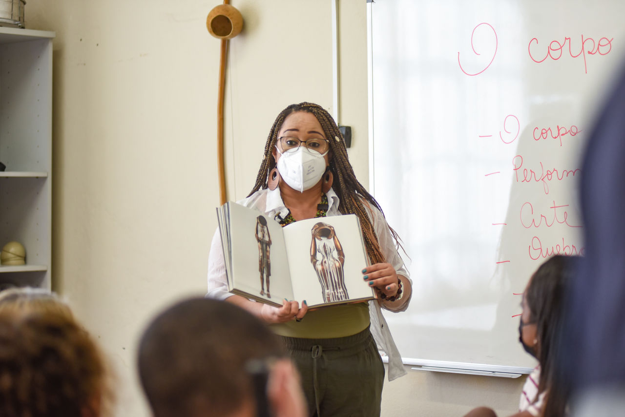 Imagem mostra mulher negra, segurando um livro aberto nas mãos, Ela é professora e está dando aula na frente de uma sala com alunos